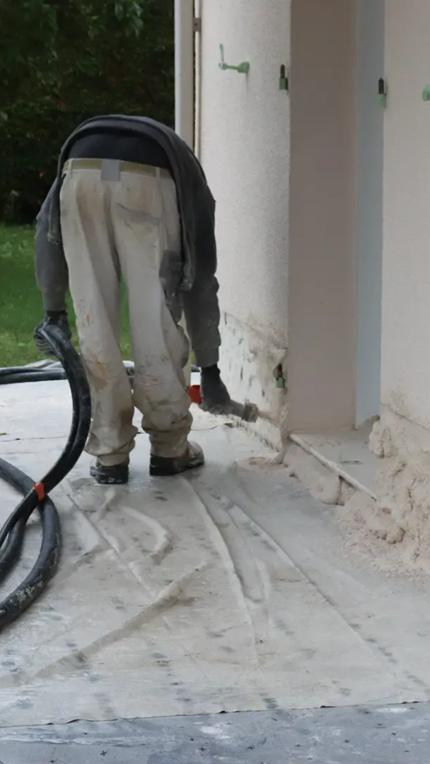 Ein Bauarbeiter trägt in einer beigen Umgebung mit Schlauch und Werkzeug Material an einer Wand auf.