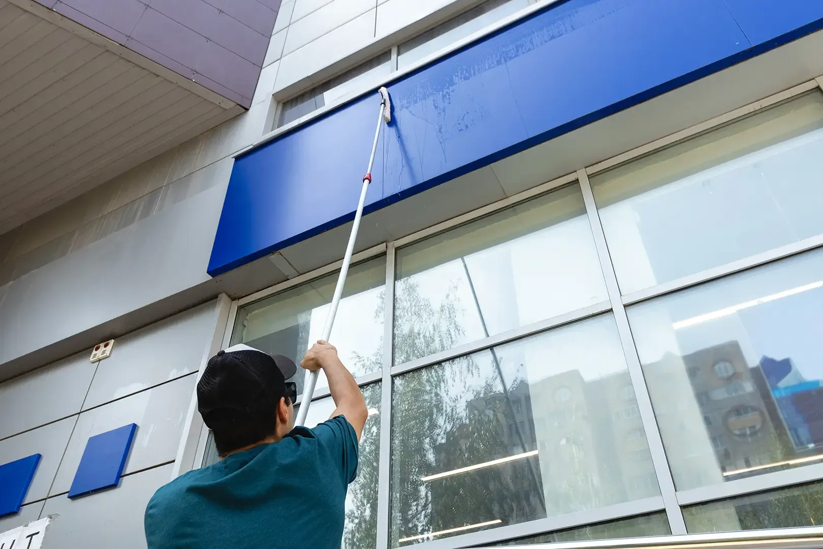 Eine Person putzt an einem sonnigen Tag mit einer Bürste mit langem Stiel die Fenster eines Gebäudes von außen.
