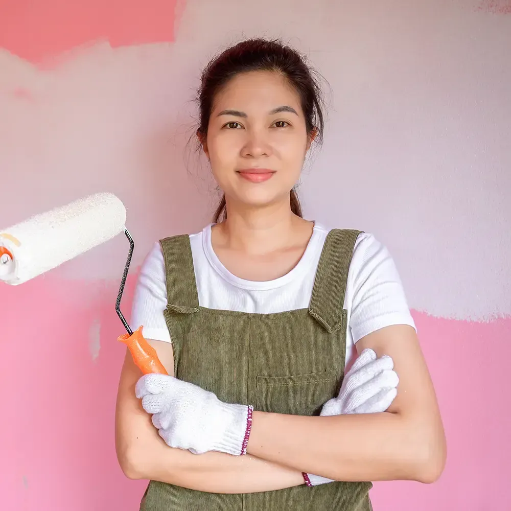 Eine Frau in Latzhose hält eine Farbrolle vor einer rosa Wand.