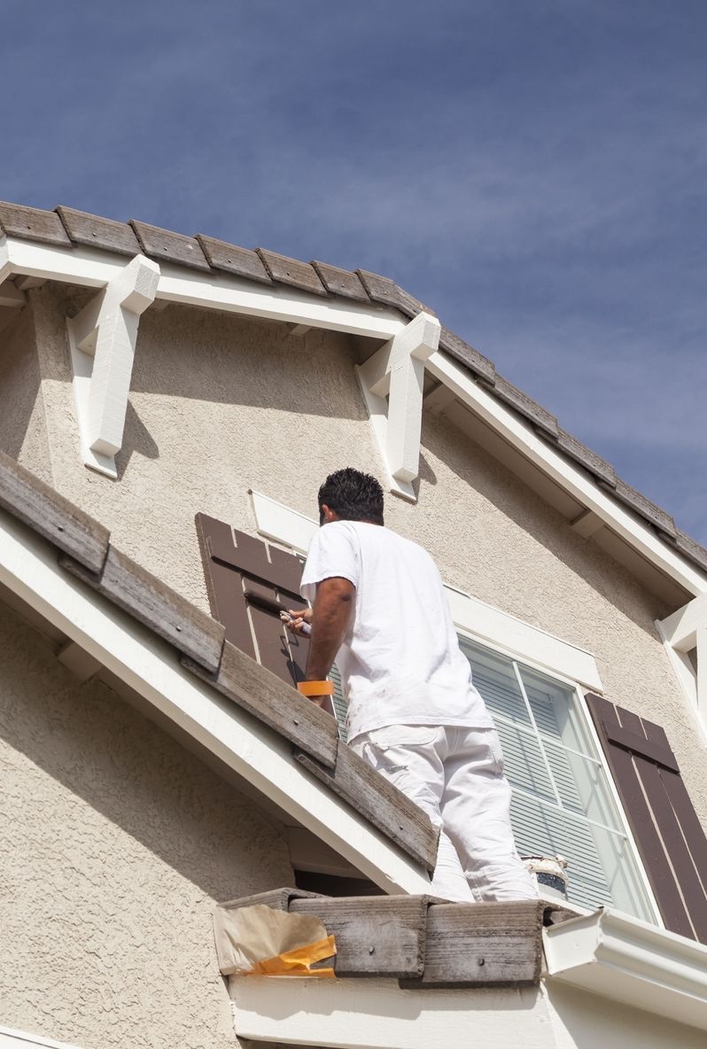 Eine Person streicht mithilfe einer Leiter und einer Farbwanne Fensterläden an einem zweistöckigen Haus.