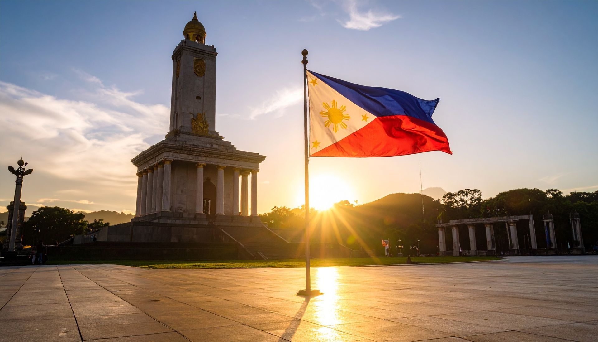 Die philippinische Flagge weht bei Sonnenuntergang in der Nähe eines Denkmals. Sonnenstrahlen strahlen über einen Platz.