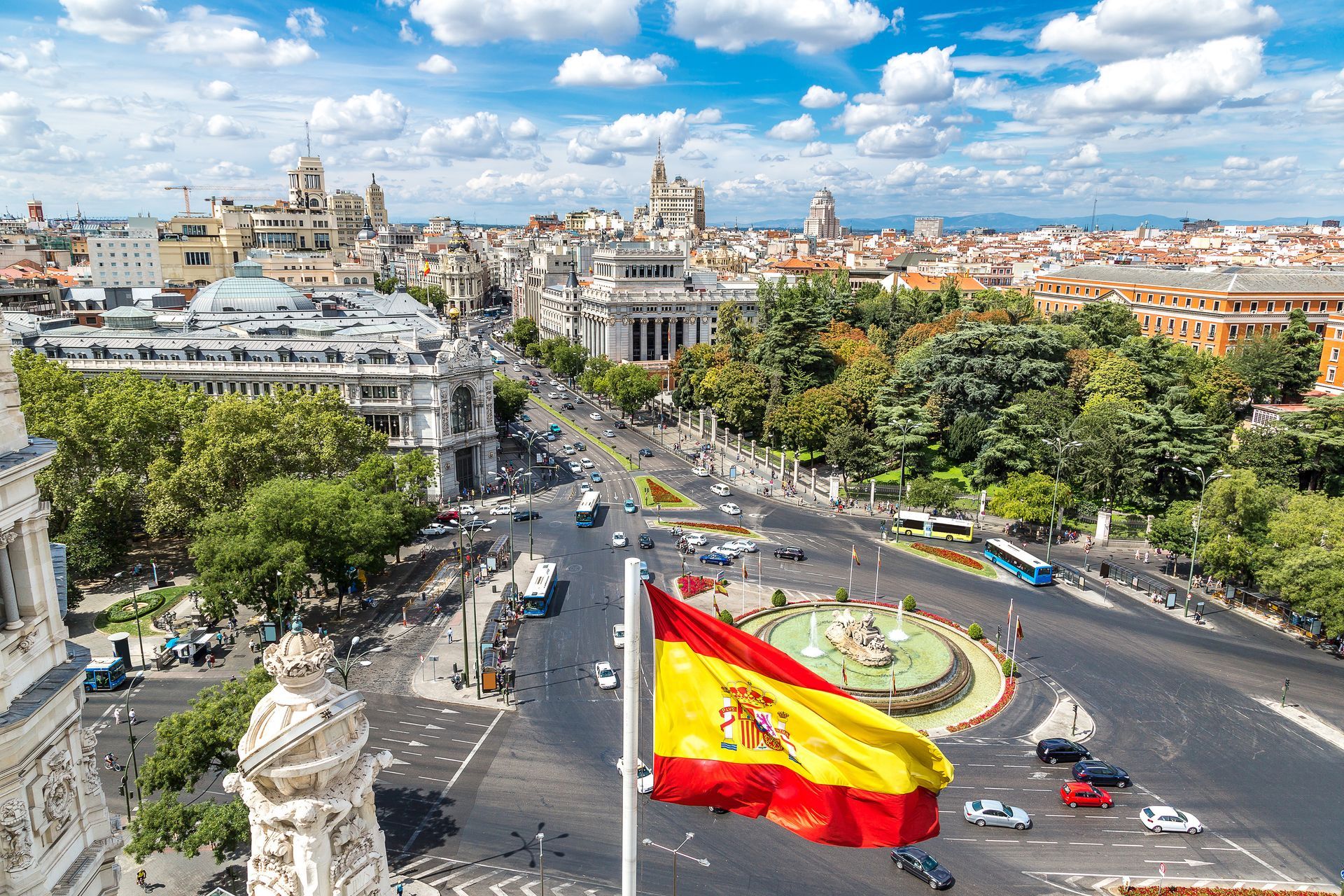 Stadtbild von Madrid mit wehender spanischer Flagge im Vordergrund; Gebäude und Verkehr im Hintergrund.