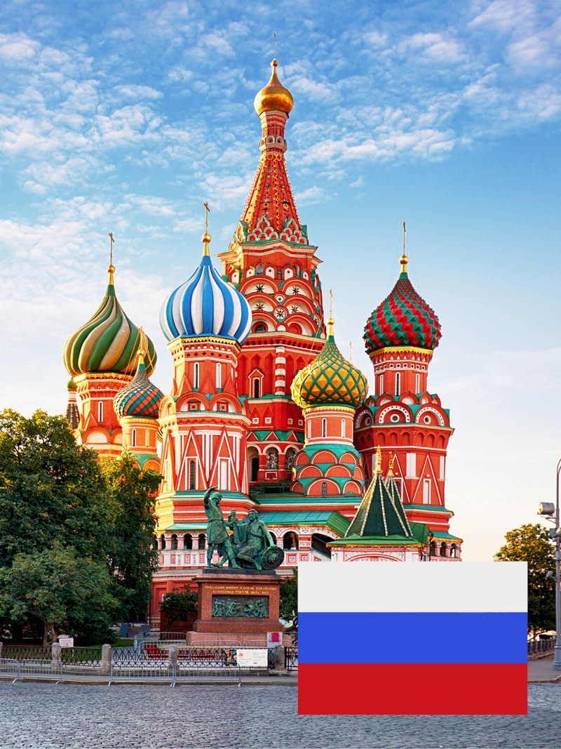 Basilius-Kathedrale in Moskau mit der russischen Flagge im Vordergrund; bunte Zwiebeltürme und rote Backsteinfassade.