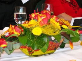 Ein farbenfrohes Blumengesteck mit gelben und roten Blüten auf einem Tisch neben einem Weinglas