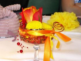 Dekoratives Blumenarrangement mit roten und gelben Blumen in einer Glasschale, mit orangefarbenem Band zusammengebunden