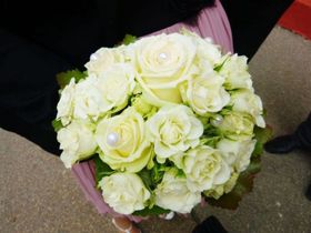 Ein Strauß weißer Rosen mit Perlenakzenten, in den Händen einer Person gehalten, vor einem Teppichhintergrund