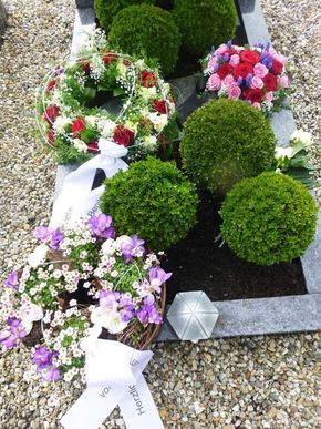 Blumengestecke mit Kränzen, Blumensträußen und grünem Formschnitt auf Kies neben einem Grabstein