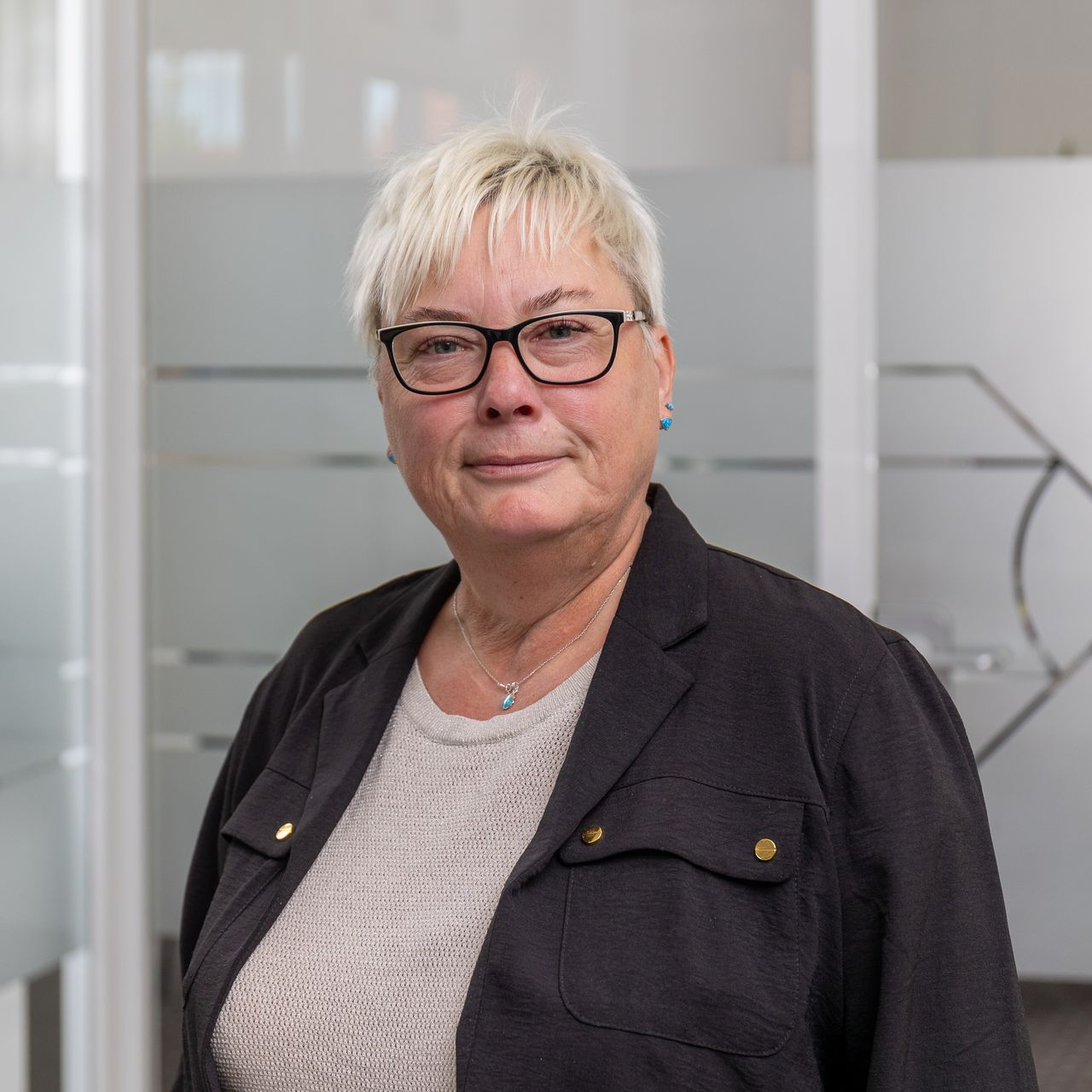 Eine Person mit kurzen Haaren, die ein weißes Oberteil mit schwarzen Punkten trägt, sitzt an einem Schreibtisch mit einem Computer in einem Büro.