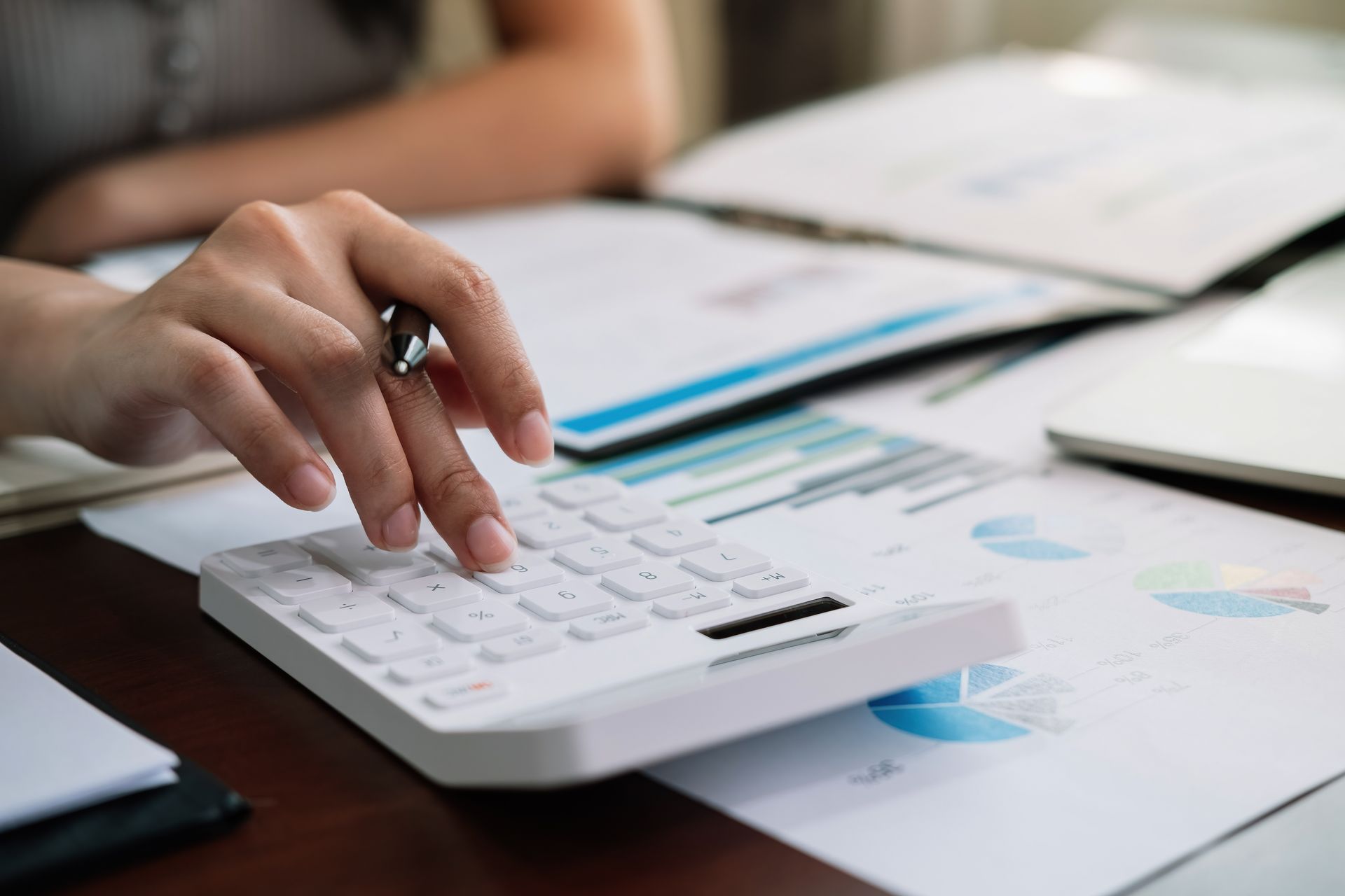 Hände, die einen weißen Taschenrechner auf einem Schreibtisch mit Finanzdiagrammen und -dokumenten bedienen.