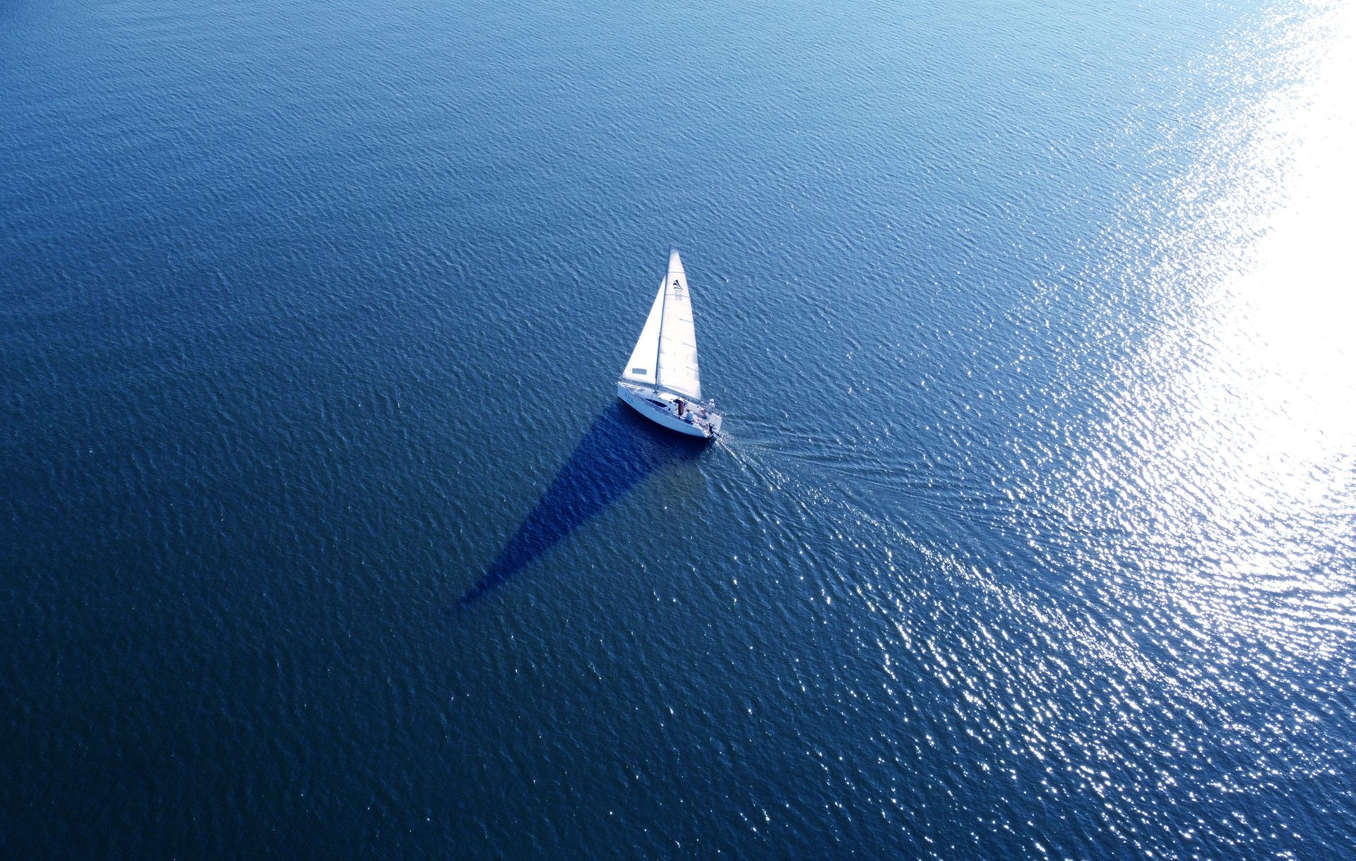 Luftaufnahme eines weißen Segelboots, das über tiefblaues Wasser fährt und einen langen Schatten wirft.