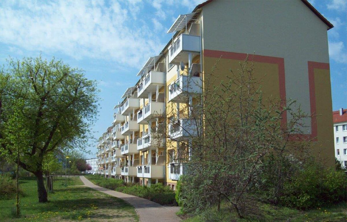 Ein mehrstöckiges Wohnhaus mit gelben und grauen Wänden und weißen Balkonen, gelegen an einem von Bäumen und Gras gesäumten Weg.