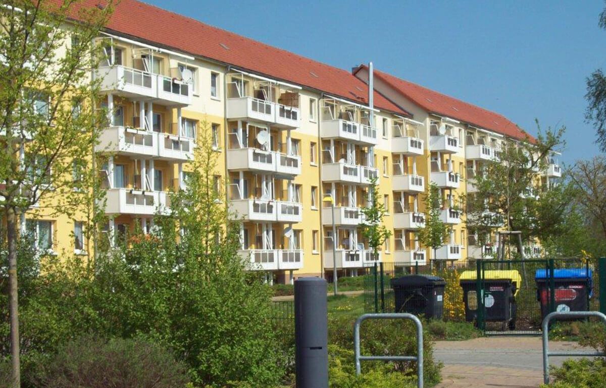 Ein mehrstöckiges Wohnhaus mit gelben Wänden, rotem Dach und weißen Balkonen, umgeben von Bäumen und Mülltonnen.