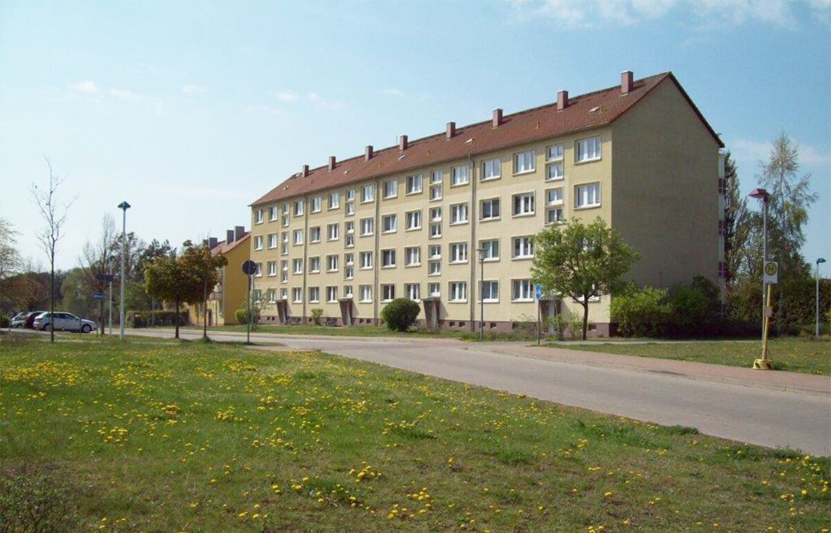 Ein mehrstöckiges, gelbes Wohnhaus mit rotem Ziegeldach, gelegen in der Nähe einer Straße und einer grünen Wiese.