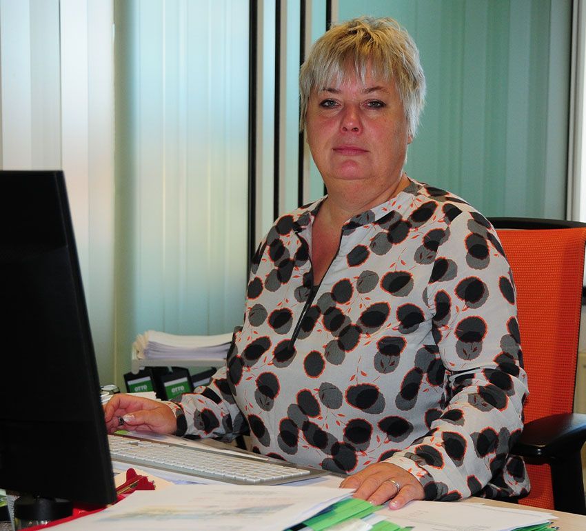 Eine Person mit kurzen Haaren, die ein weißes Oberteil mit schwarzen Punkten trägt, sitzt an einem Schreibtisch mit einem Computer in einem Büro.