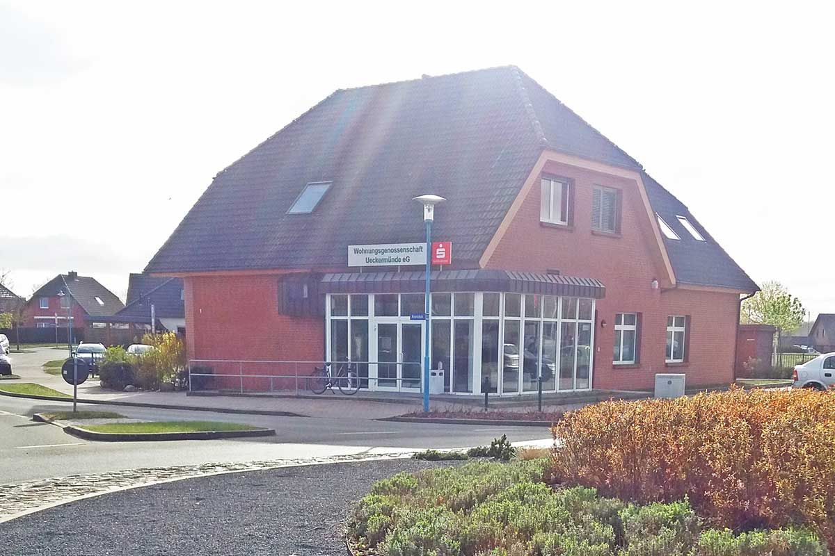 Ein rotes Backsteingebäude mit großem Glaseingang, Dachgauben und steilem Dach, gesehen von einem Eckgrundstück aus.