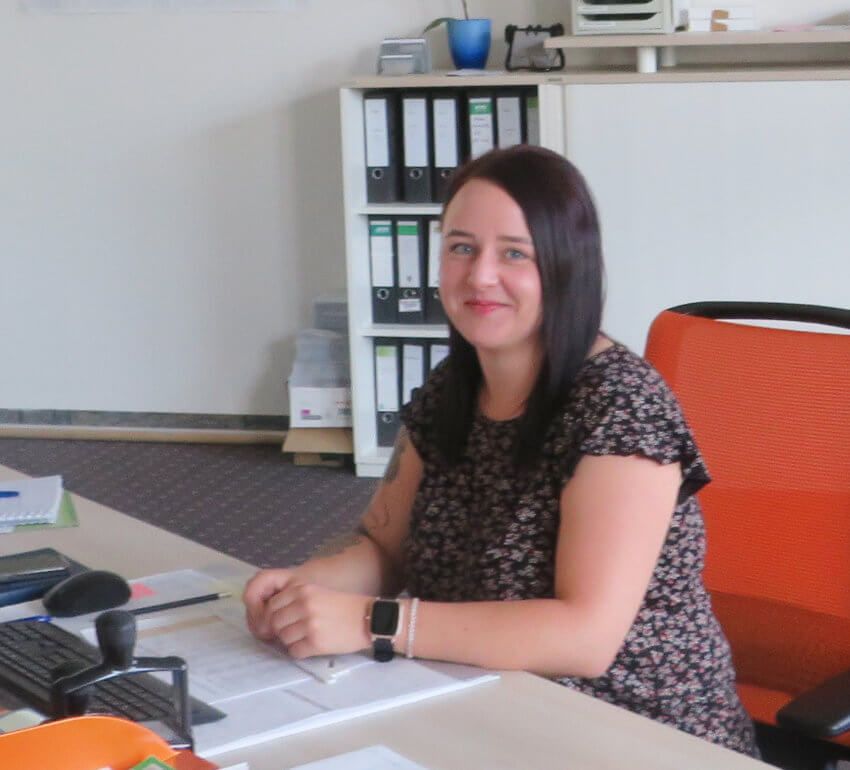 Eine Person mit dunklen Haaren sitzt an einem Schreibtisch im Büro und lächelt in die Kamera. Neben dem Schreibtisch stehen Regale voller Ordner und ein orangefarbener Stuhl.