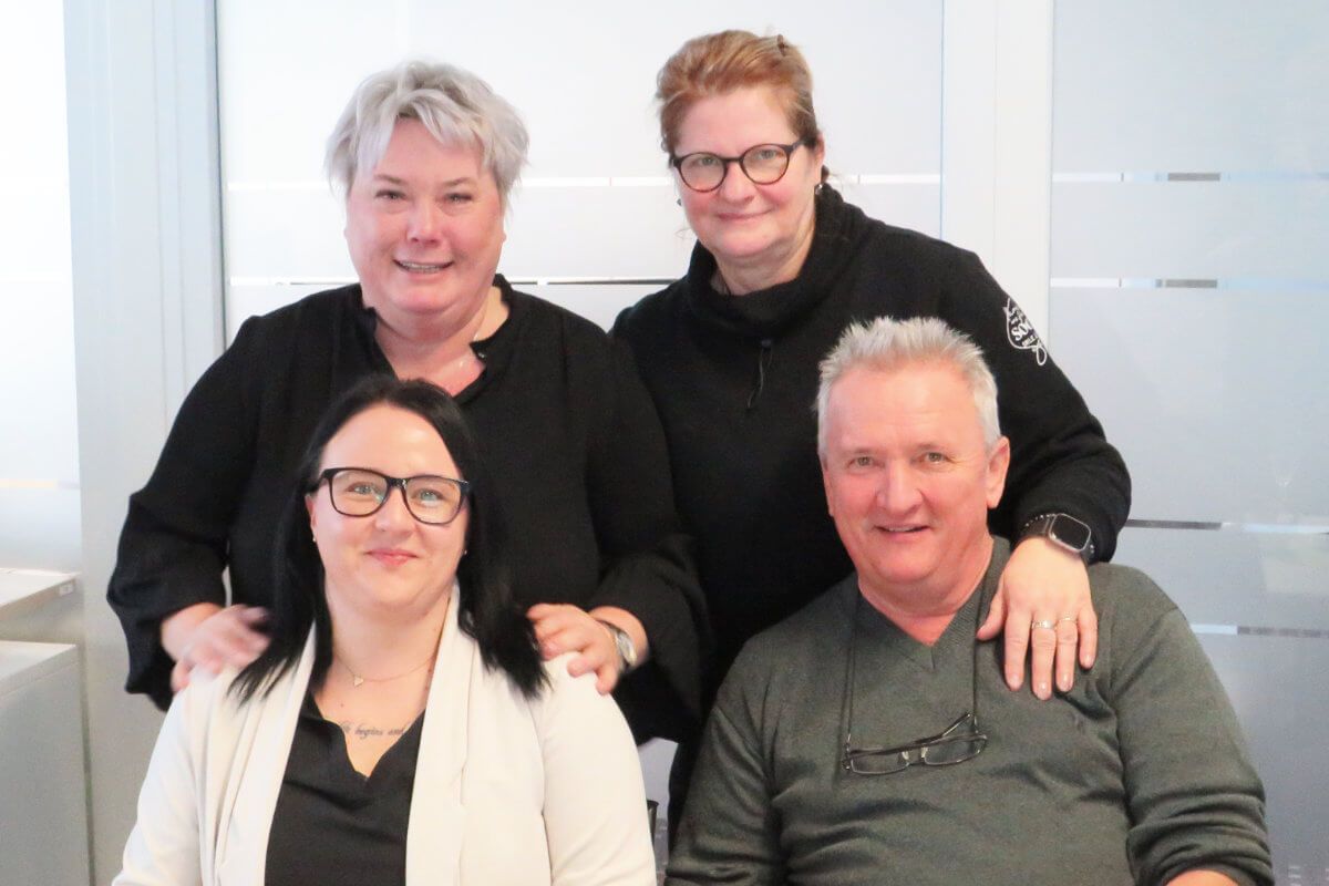 Zwei Frauen stehen hinter einer anderen Frau und einem Mann, alle lächeln, und posieren gemeinsam in einer Büroumgebung.