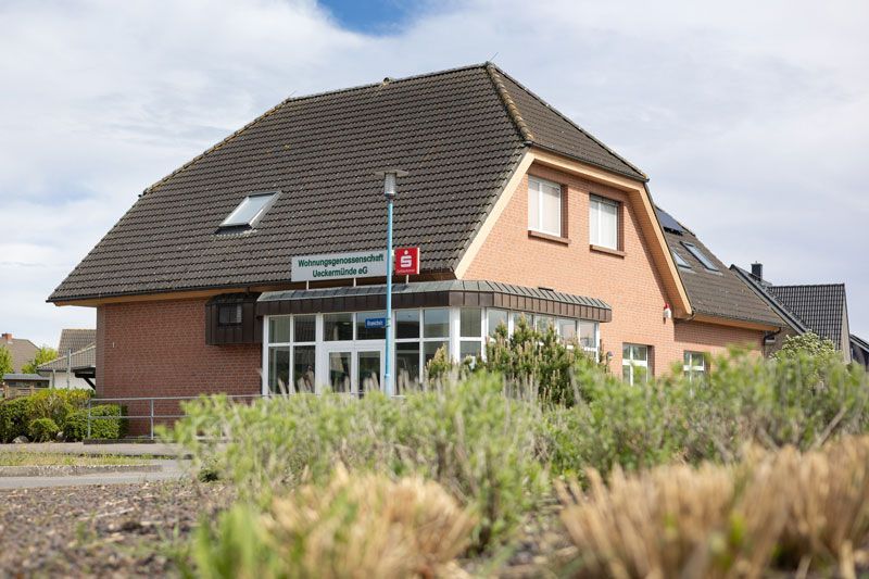 Ein rotes Backsteingebäude mit großem Glaseingang, Dachgauben und steilem Dach, gesehen von einem Eckgrundstück aus.