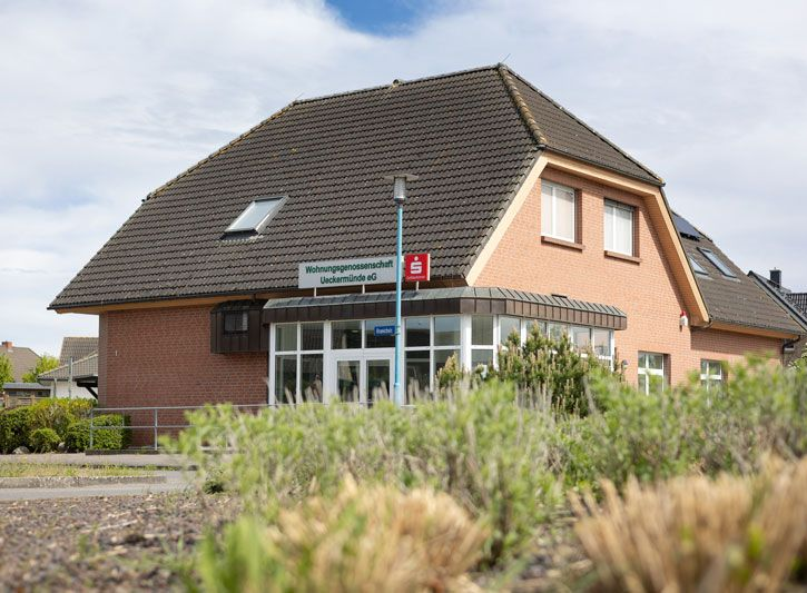 Ein rotes Backsteingebäude mit großem Glaseingang, Dachgauben und steilem Dach, gesehen von einem Eckgrundstück aus.