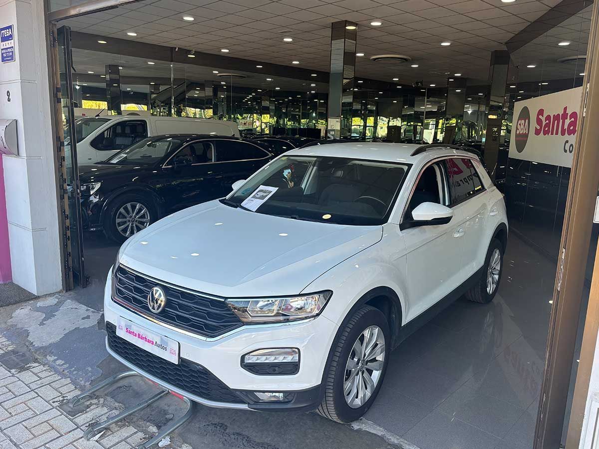 SUV Volkswagen T-Roc blanco estacionado dentro de un concesionario de automóviles, con otros vehículos visibles al fondo.