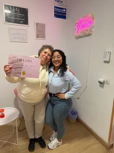 Dos mujeres sonríen en una habitación; una de ellas sostiene un papel 