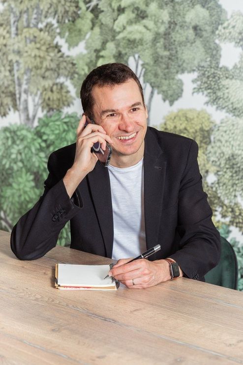 Un homme en blazer noir, assis à une table, tient un téléphone à l'oreille et sourit.