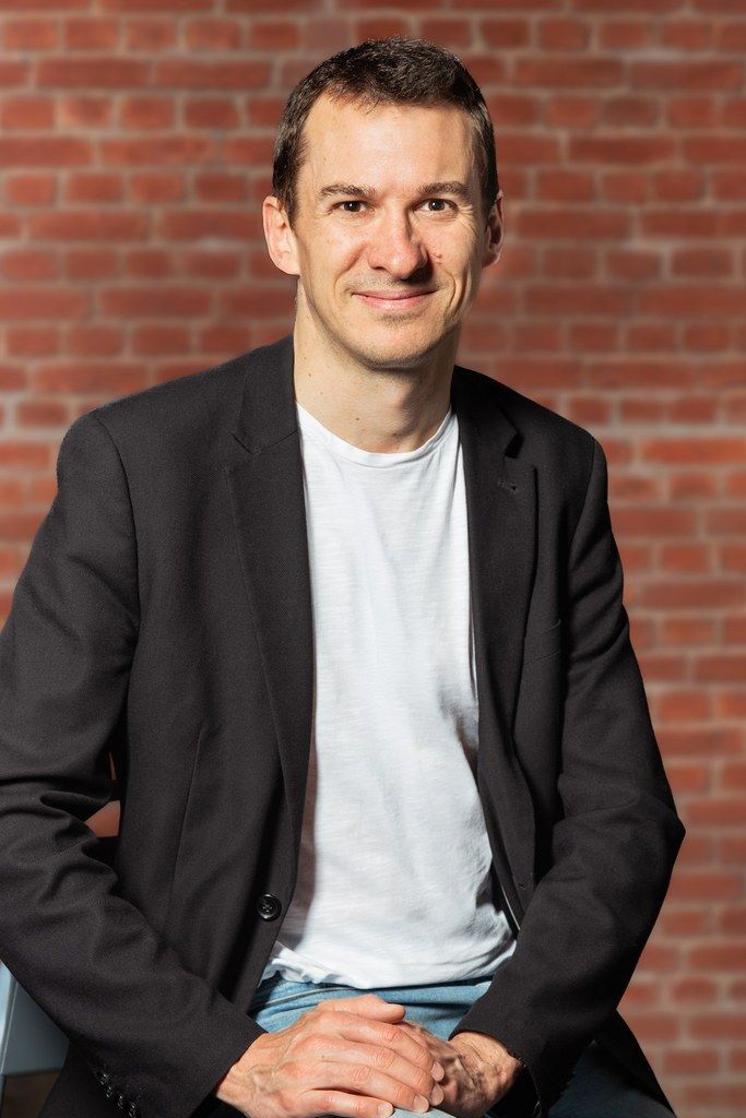 Un homme assis, vêtu d'un blazer noir et d'une chemise blanche, est adossé à un mur de briques et esquisse un sourire.