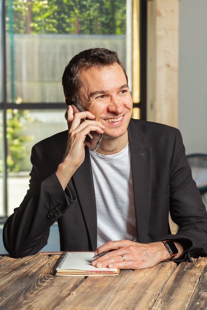 Un homme souriant parle au téléphone à une table en bois dans un bureau lumineux