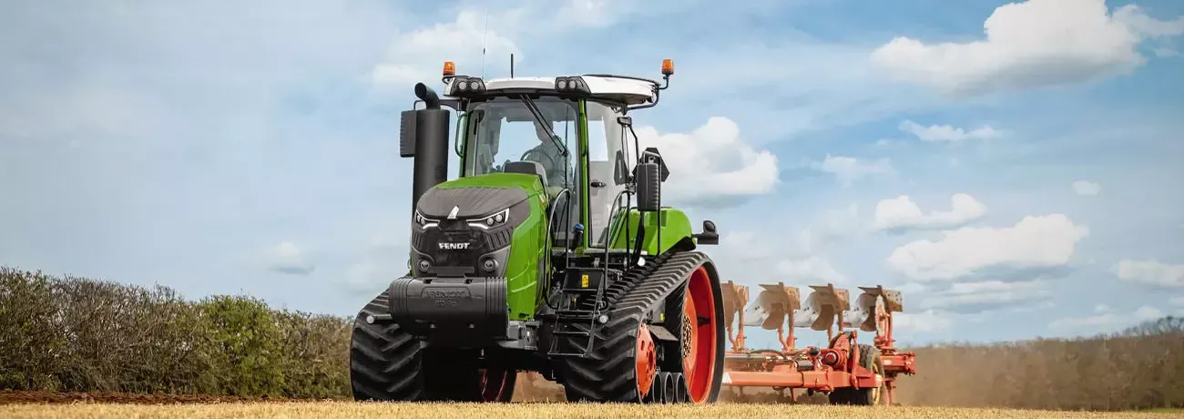 Un tractor ara un campo bajo un cielo azul nublado, con aperos agrícolas naranjas enganchados en la parte trasera.