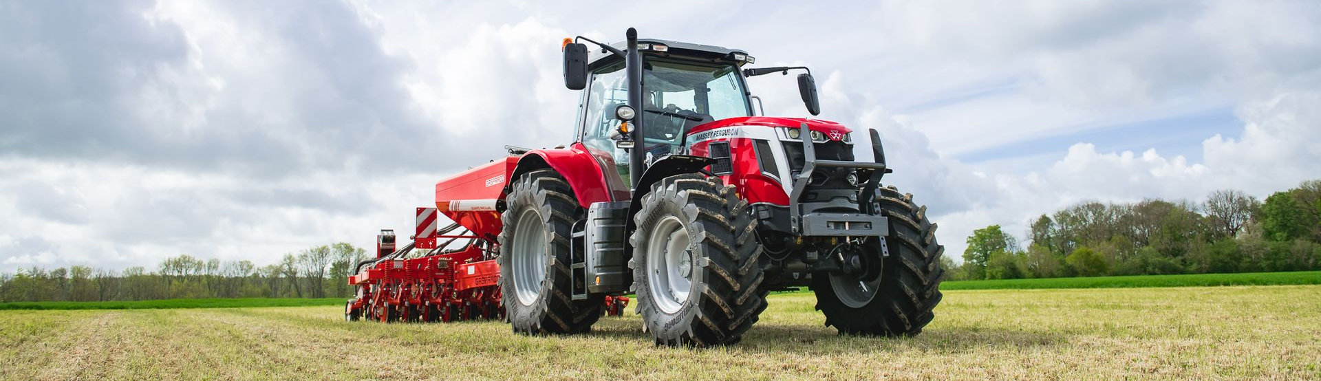 Un tractor rojo arrastrando maquinaria agrícola a través de un campo de hierba bajo un cielo nublado.