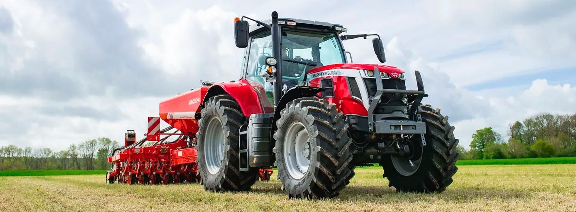 Un tractor rojo arrastrando maquinaria agrícola a través de un campo verde bajo un cielo nublado.