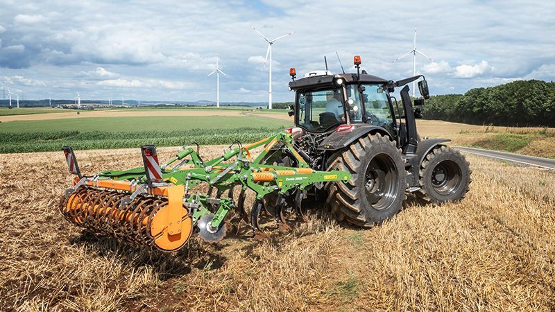 Un tractor arrastrando un implemento agrícola a través de un campo cosechado bajo un cielo nublado con aerogeneradores en la distancia.