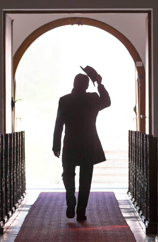 Silhouette eines Mannes im Mantel, der seinen Hut lüftet und durch einen Torbogen auf ein helles Licht zugeht.