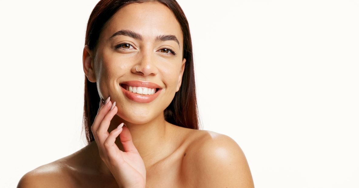 Mujer de cabello y piel castaños sonríe, tocándose el cuello contra un fondo blanco.