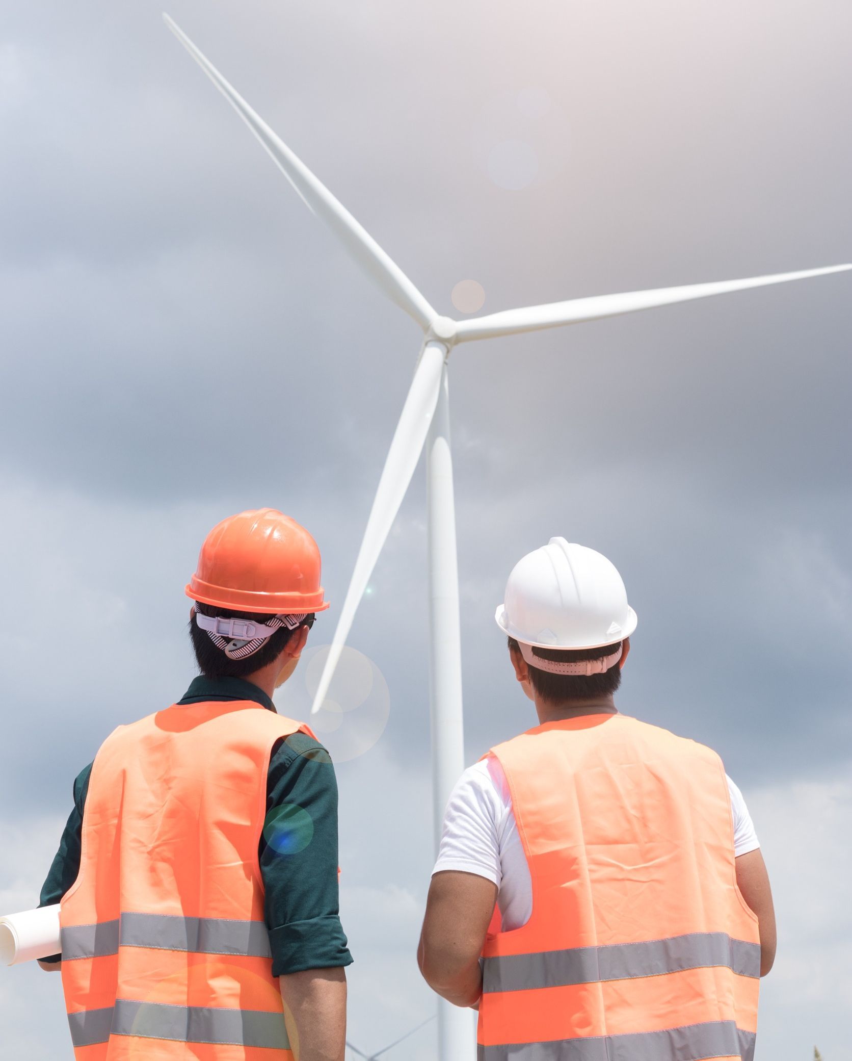 Dos hombres con chalecos naranjas y cascos miran una turbina eólica.