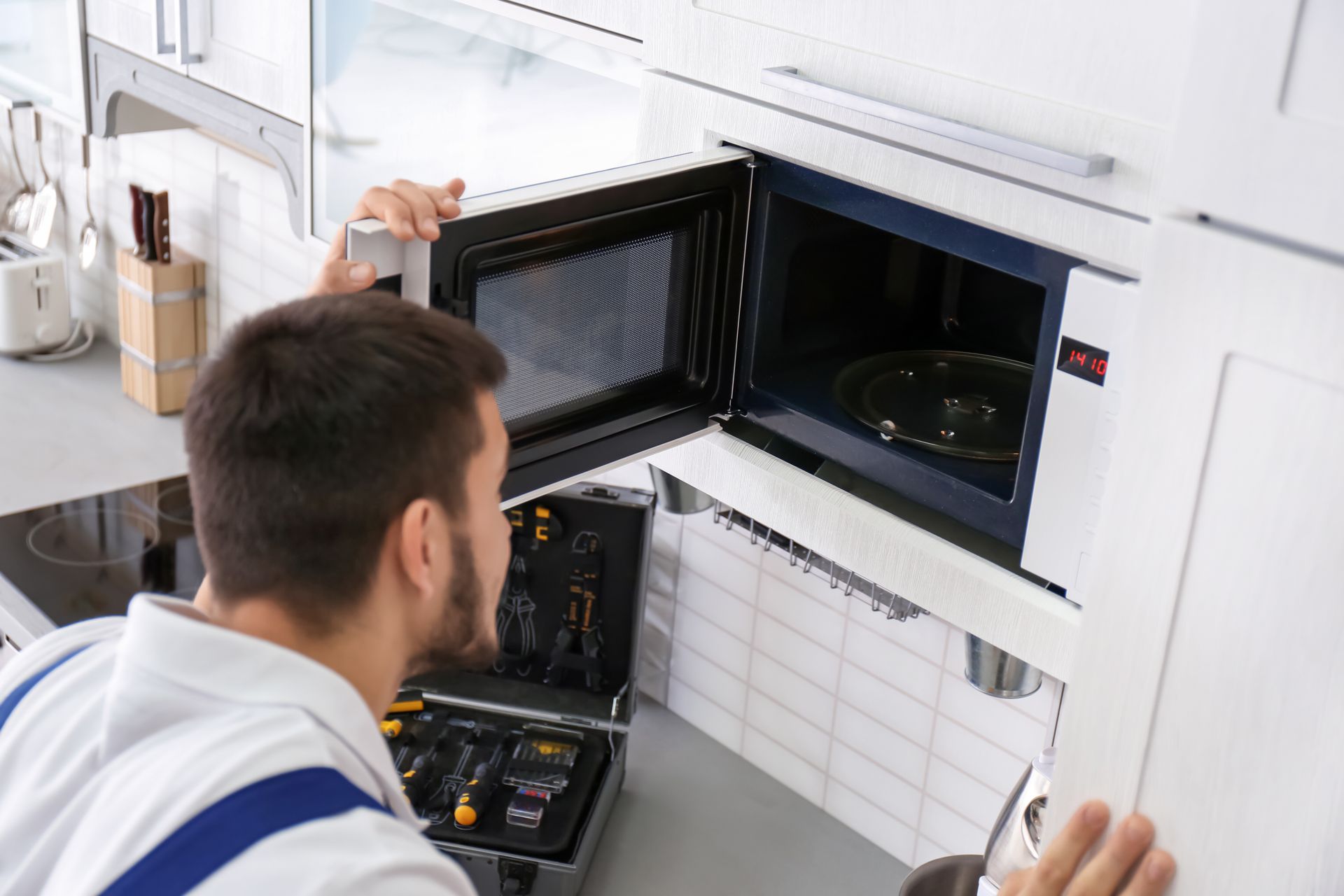 Un technicien en uniforme blanc inspecte un four à micro-ondes encastré et ouvert dans une cuisine moderne carrelée de blanc.