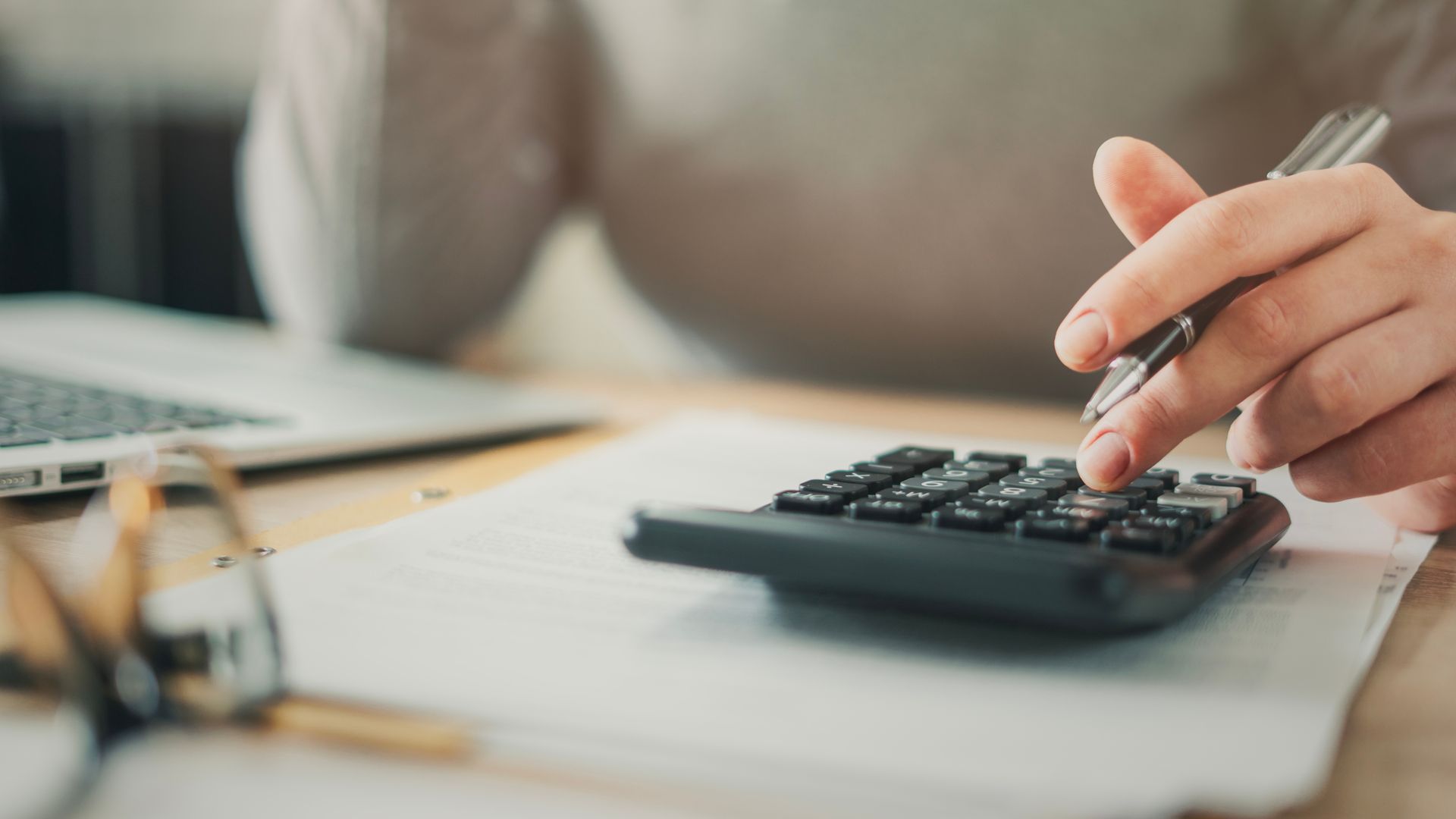 Une personne utilise un stylo pour manipuler une calculatrice à côté de documents et d'un ordinateur portable posés sur un bureau en bois.