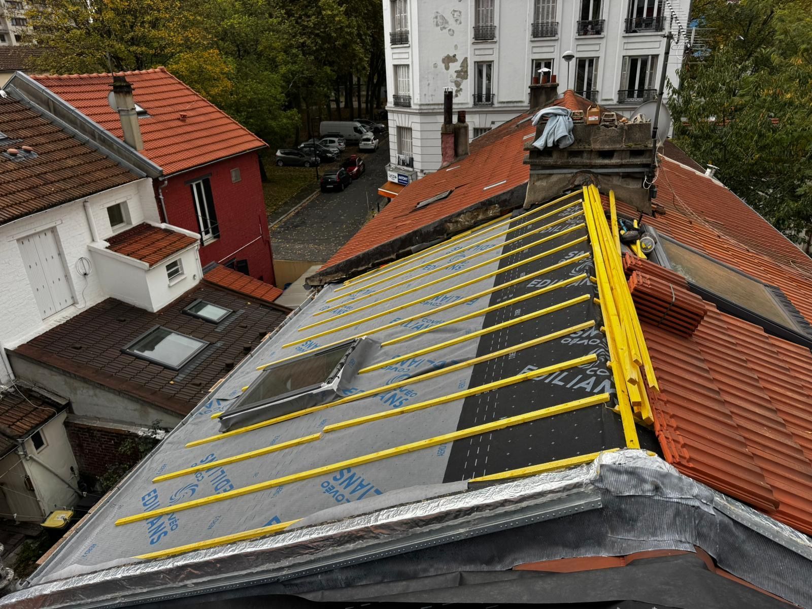 Toiture avec puits de lumière, cheminée et matériaux de couverture neufs. Tuiles orange et grises, supports jaunes.