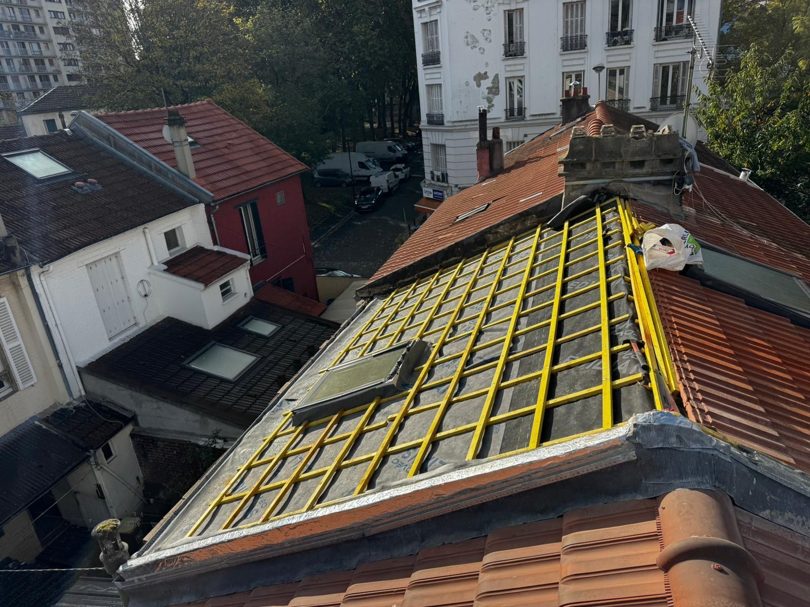 Vue de toits, avec un toit partiellement rénové présentant une charpente jaune et des puits de lumière, en milieu urbain.
