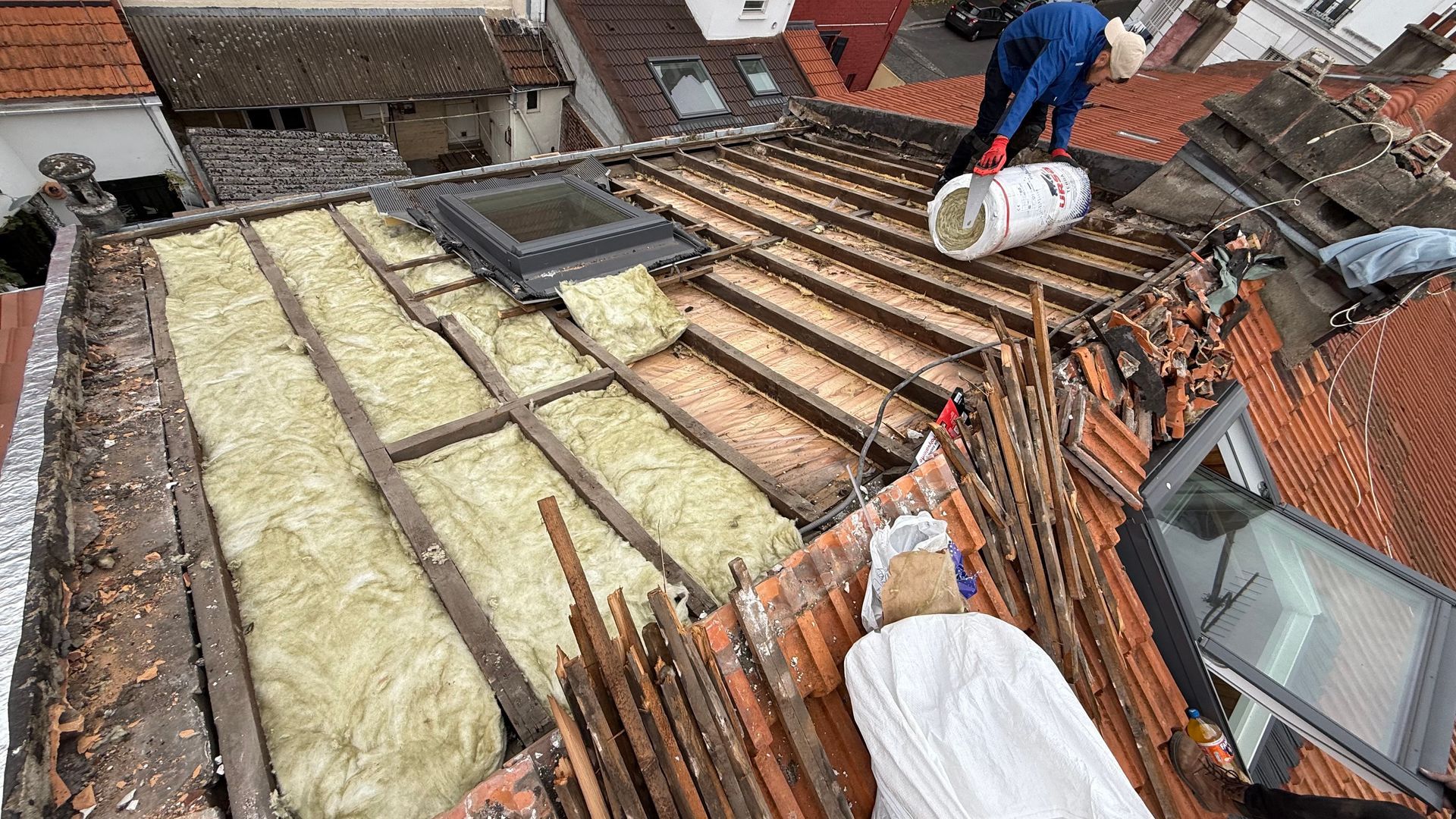 Pose d'isolant de toiture : Un ouvrier sur un toit de tuiles rouges installe de l'isolant entre des poutres en bois 