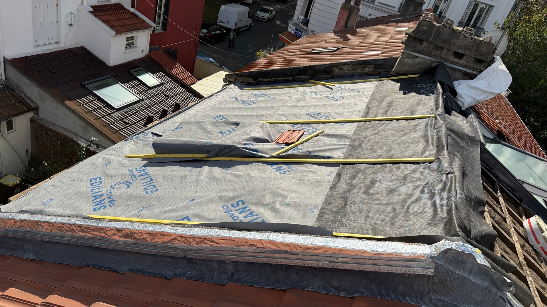 Toiture partiellement recouverte d'un matériau imperméable gris, guides et outils jaunes, surplombant les bâtiments.