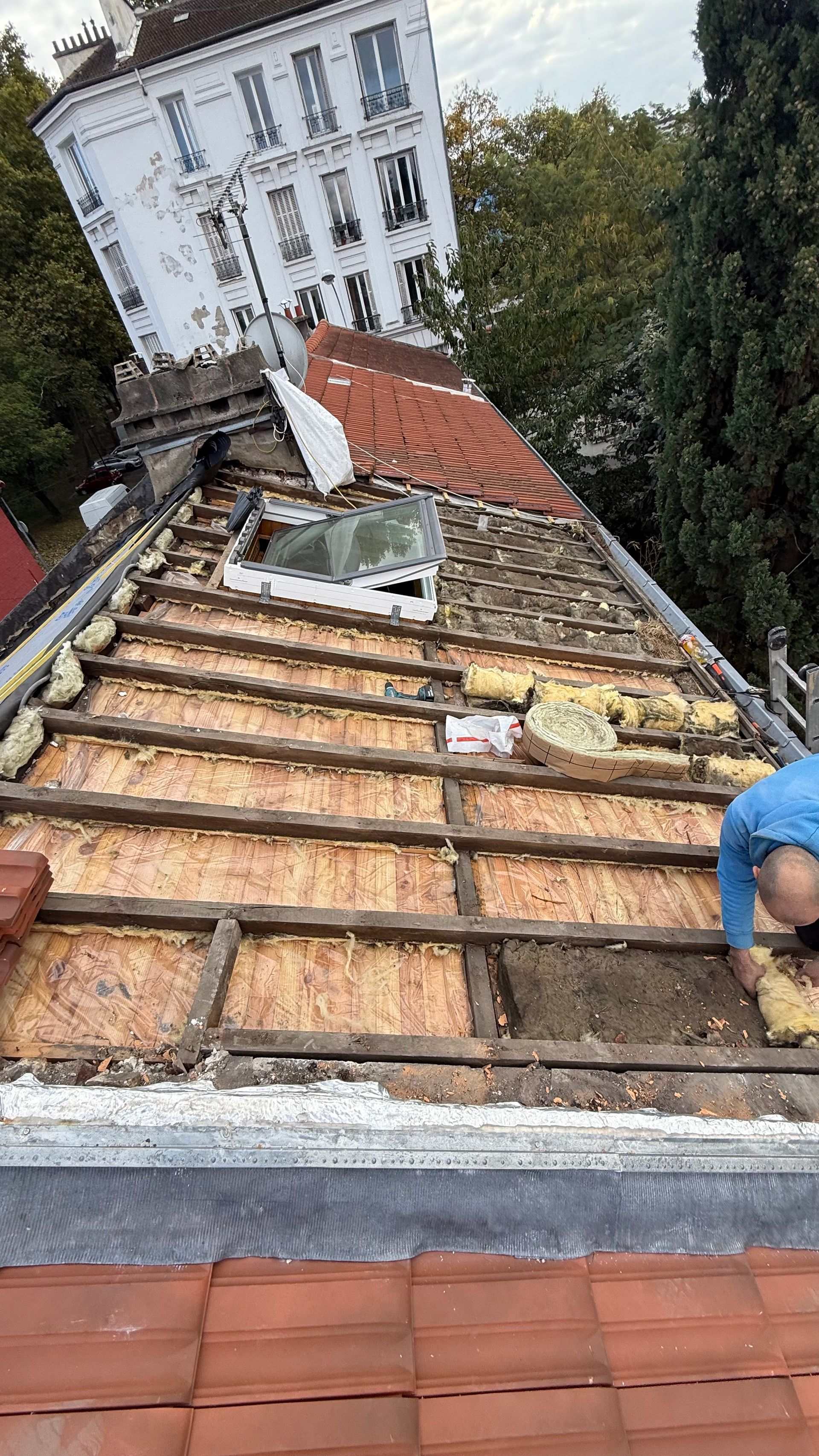 Toiture en construction, charpente apparente, ouvrier à l'œuvre. Un bâtiment blanc se dresse en arrière-plan.