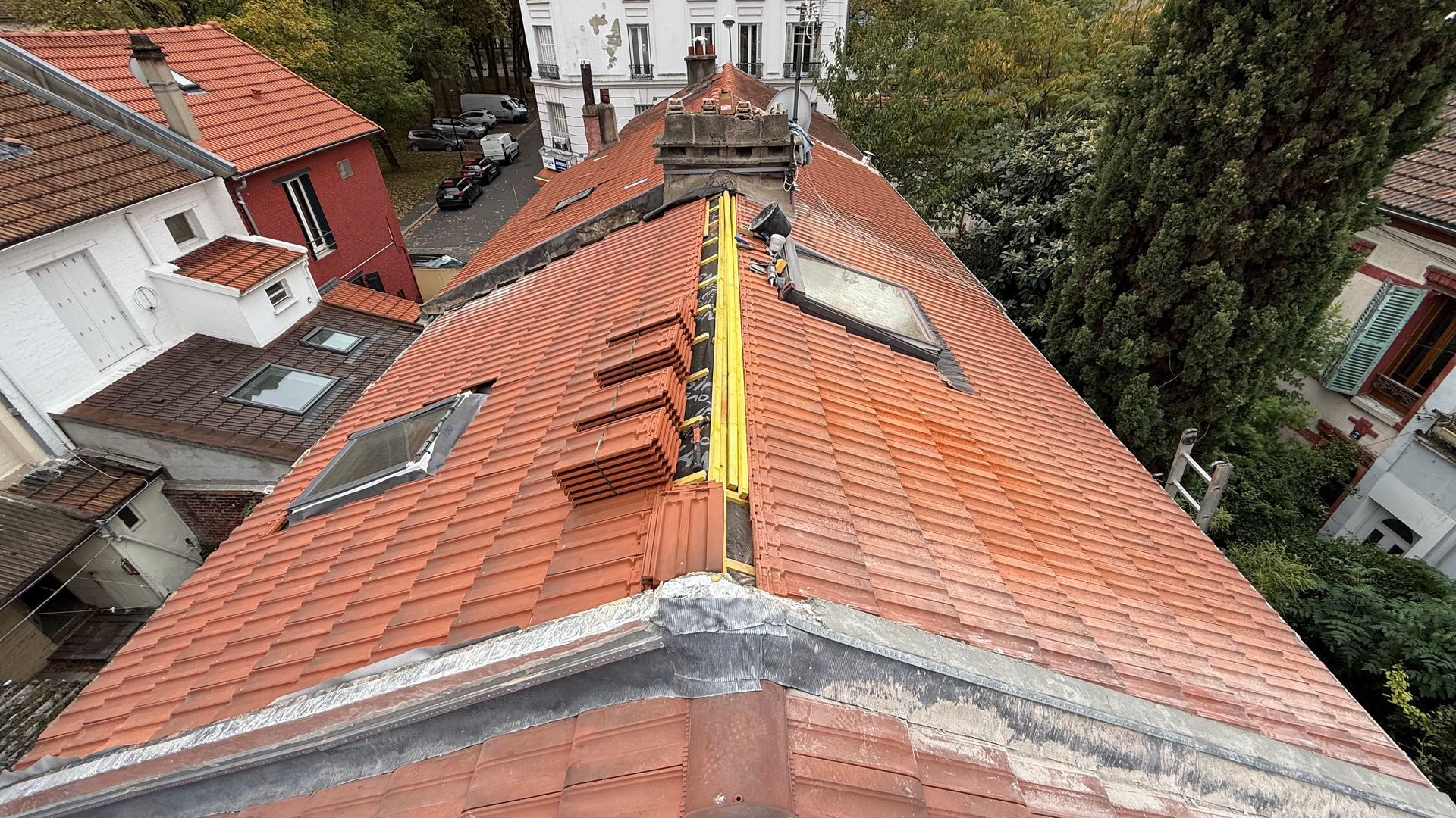 Toit en tuiles rouges avec puits de lumière, cheminées et bâtiments environnants.