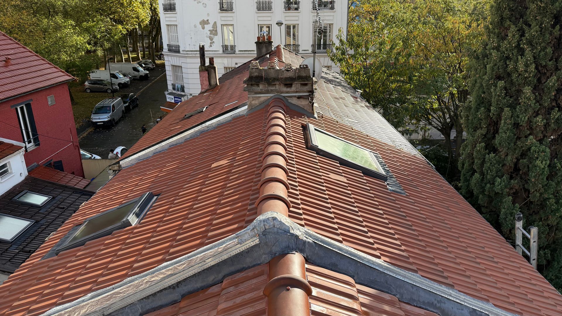 Vue d'un toit de tuiles en terre cuite avec puits de lumière, cheminée et bâtiments environnants sous un ciel ensoleillé.