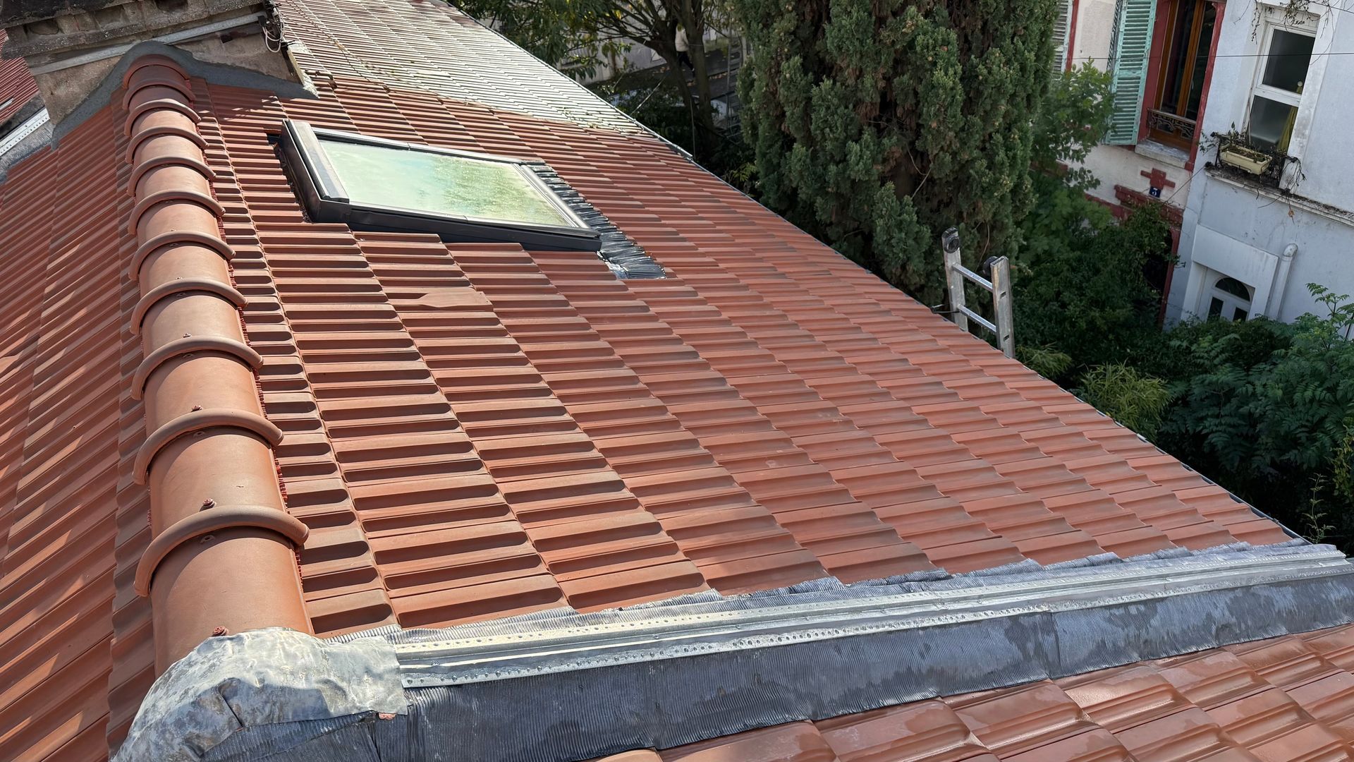 Toit en tuiles rouges avec puits de lumière ; vue depuis le toit avec un bâtiment et des arbres en arrière-plan.