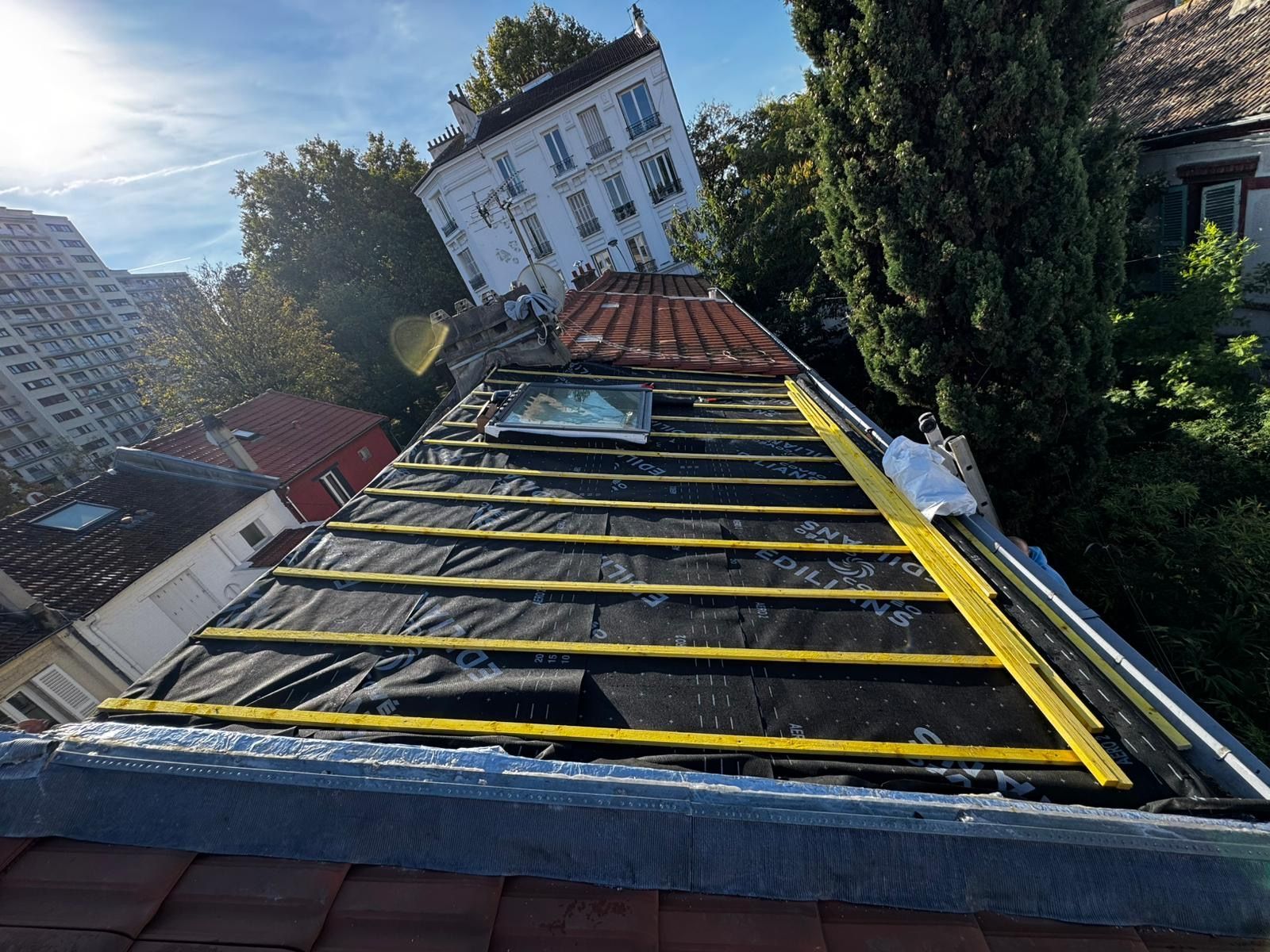 Toiture avec sous-couche noire, liteaux jaunes, puits de lumière et outils ; bâtiments et arbres en arrière-plan.