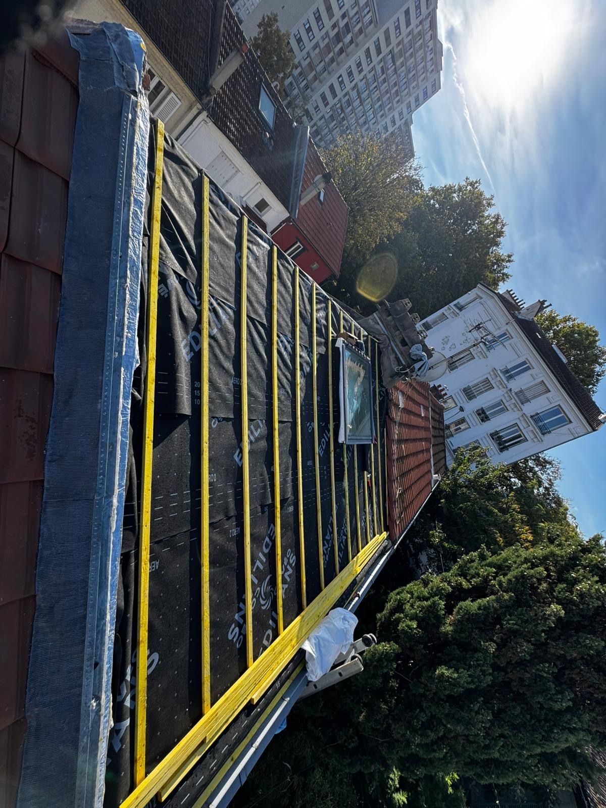 Construction sur le toit avec sous-couche noire, liteaux jaunes et bâtiments environnants sous un ciel ensoleillé.