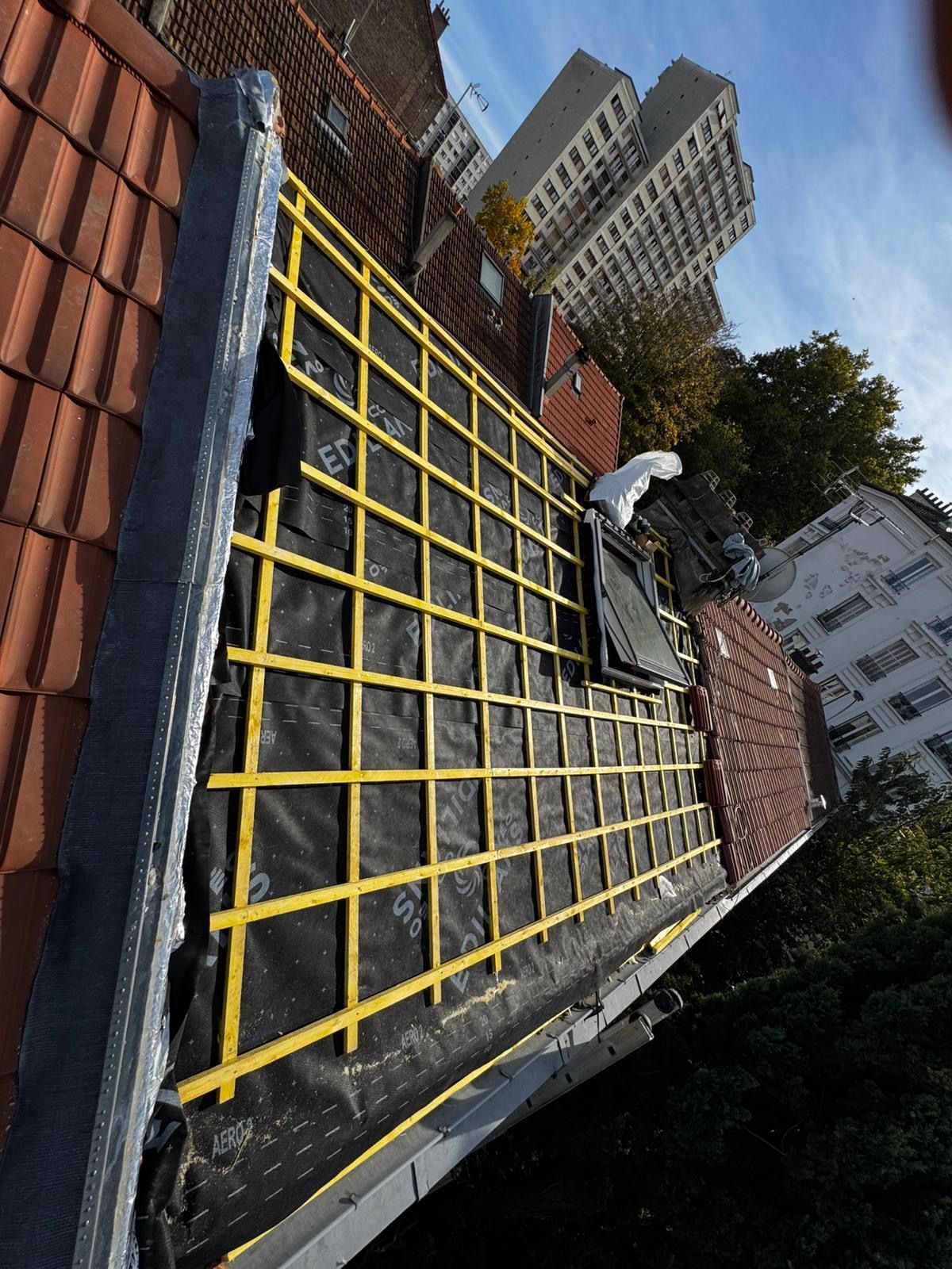 Travaux de réparation de toiture en cours ; grille jaune sur sous-couche noire. Tuiles et briques rouges visibles.