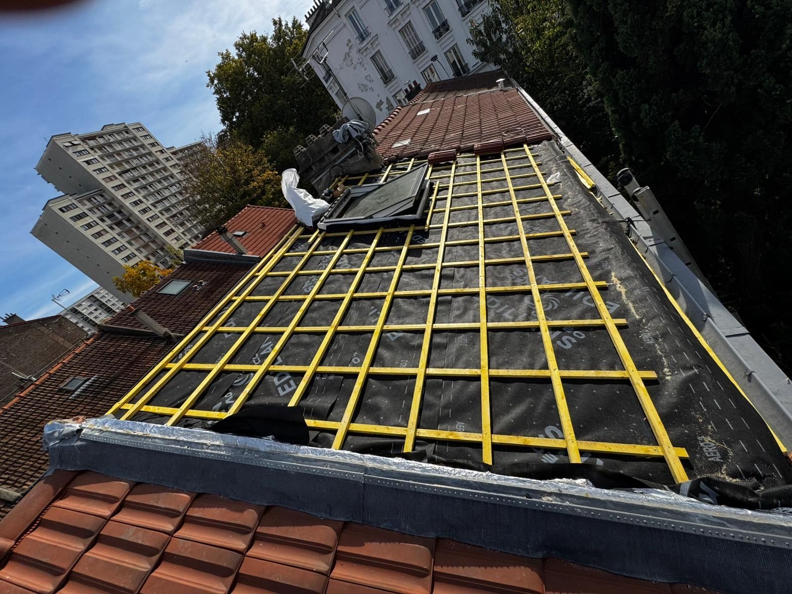 Toiture en construction ; bâche noire recouverte d'un quadrillage jaune, tuiles rouges en dessous, bâtiments en arrière-plan.