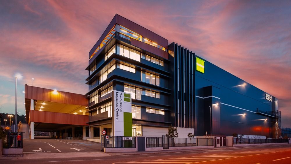 Moderno edificio de oficinas al anochecer, con fachada oscura, ventanas iluminadas y un cielo colorido. Un cartel indica el nombre del edificio en verde y blanco.